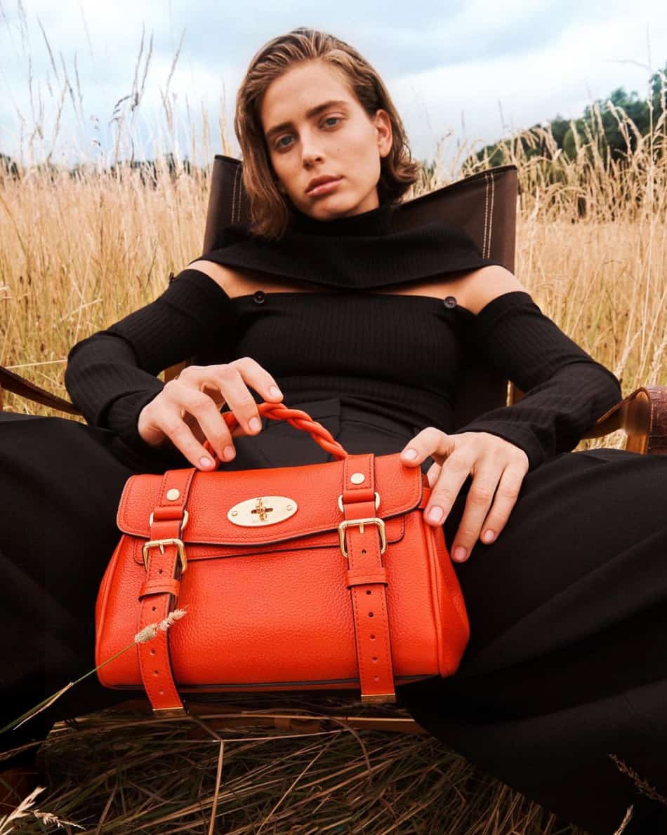 femme se penchant en arrière sur une chaise vêtue de noir et tenant un sac Mulberry rouge