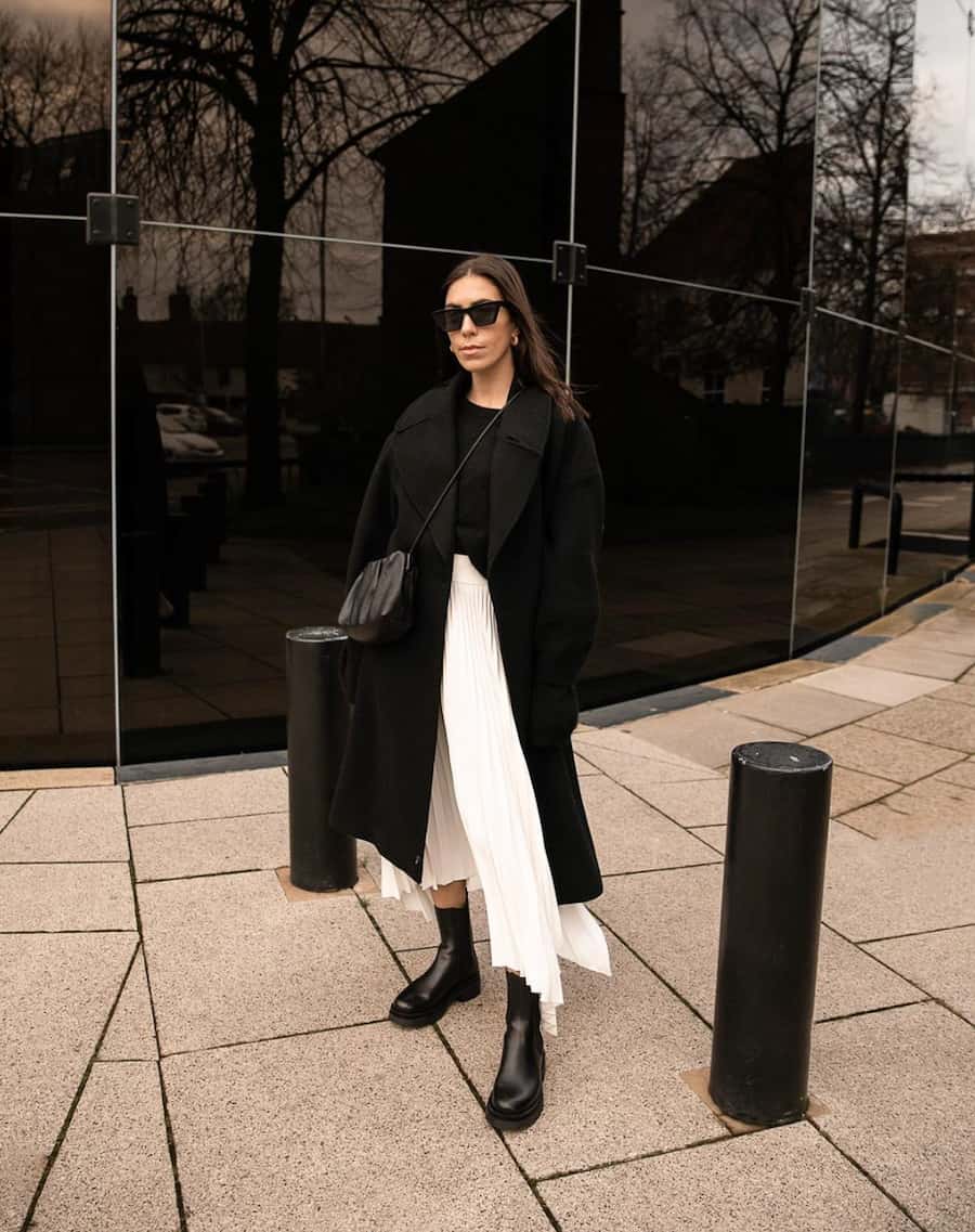 femme portant un long manteau noir avec une jupe plissée ivoire et des bottes noires