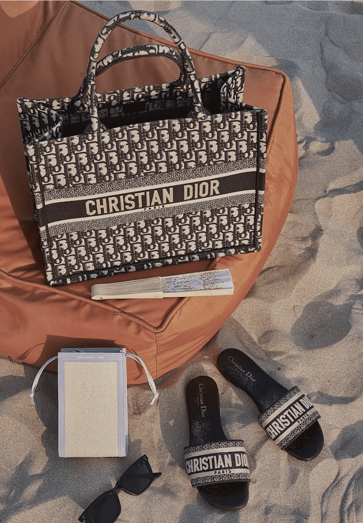image d'un sac fourre-tout Dior posé sur un pouf sur la plage avec des sandales et des lunettes de soleil Dior à côté