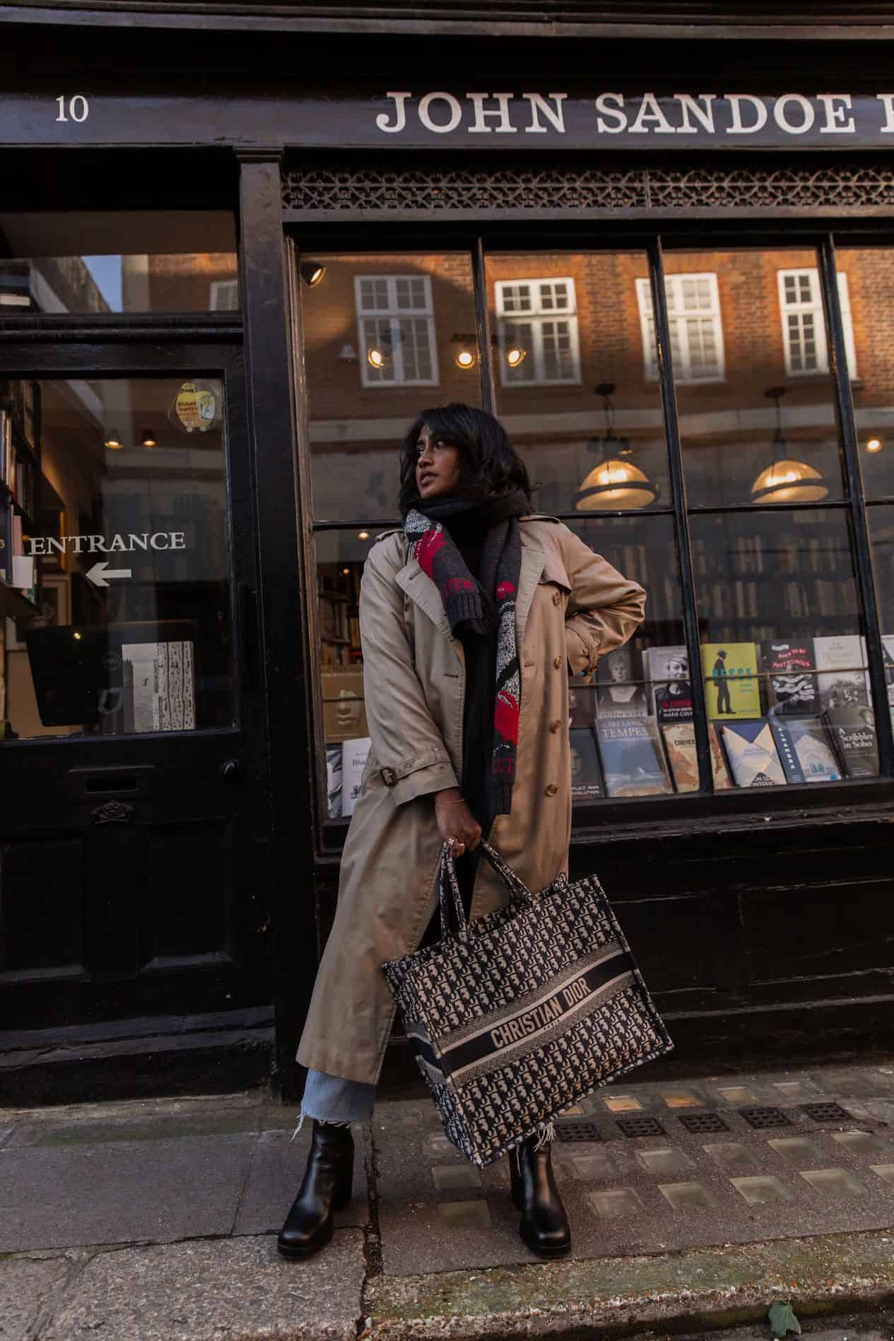 image d'une femme brune debout devant une librairie portant un long trench-coat beige et tenant un fourre-tout Dior